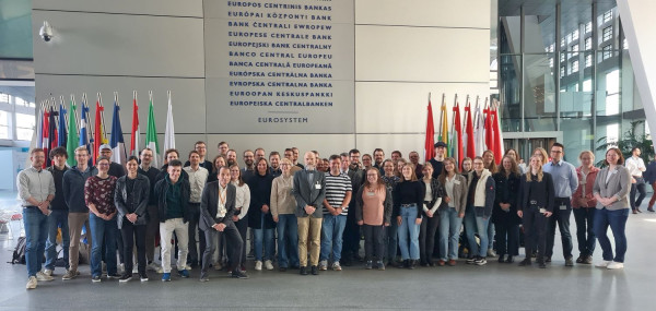 Eine Gruppe Menschen steht vor aufgestellten Fahnen in einem Gebäude