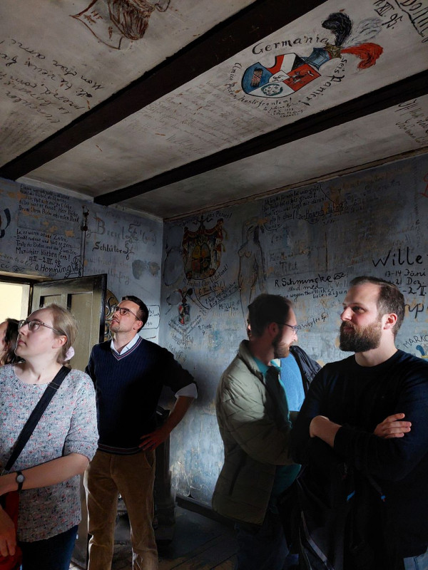 Ein paar Menschen stehen in einem halbdunklen Raum. An Wand und Decke sind Inschriften und Malereien zu sehen.