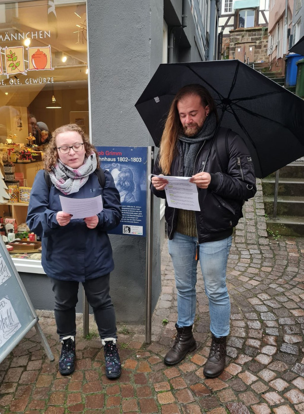 Zwei Menschen stehen im Freien vor einem Schaufenster beziehungsweise vor der Einmündung einer Gasse und halten Zettel in der Hand. Eine der Personen hat einen Regenschirm aufgespannt.