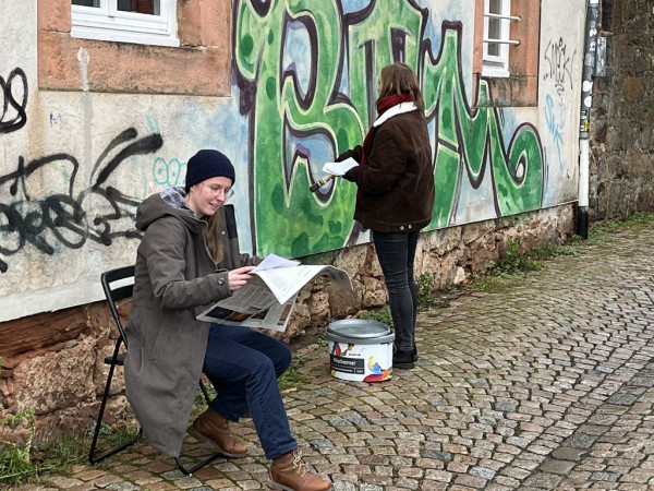 Eine Frau sitzt im Freien auf einem Stuhl und hält Zettel in der Hand. Im Hintergrund steht ein Gebäude mit Graffitis, an dem eine andere Frau malt.