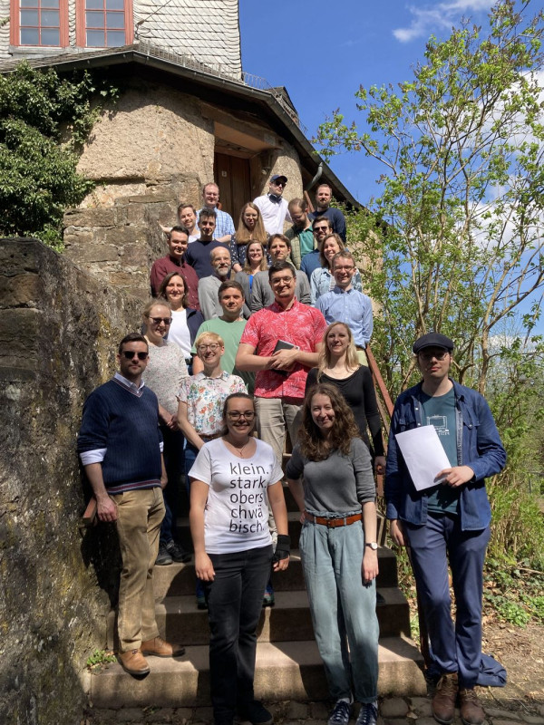 Eine Gruppe Menschen steht im Freien auf einer Treppe. Seitlich sind Mauern und ein Gebäude zu sehen.