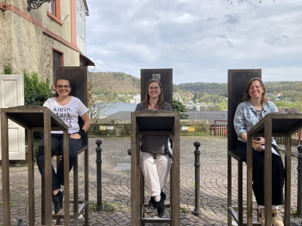 Drei Frauen sitzen im Freien auf hohen Stühlen. Vor ihnen stehen Pulte.
