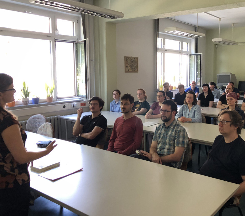In einem Raum mit geöffneten Fenstern sitzen Menschen in mehreren Reihen hinter Tischen. Vorne steht eine Frau und spricht.