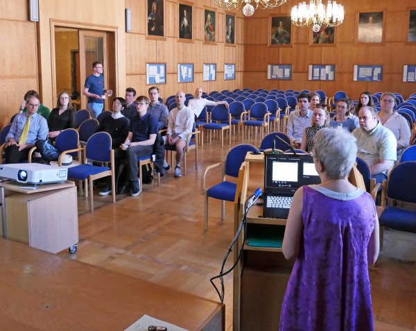 Mehrere Menschen sitzen in einem historischen Saal, im Vordergrund steht eine Frau und trägt vor.