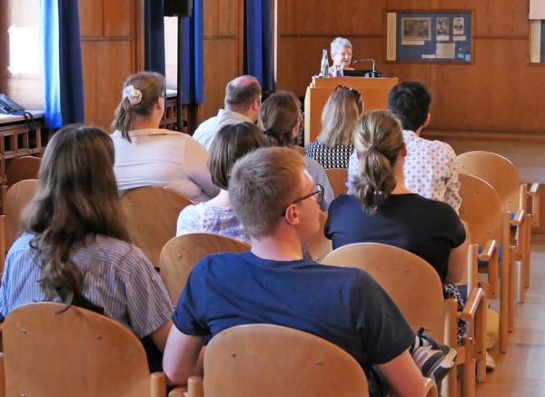 Im Vordergrund sitzen Menschen in einem Saal. Im Hintergrund steht am Pult eine Frau und trägt vor.