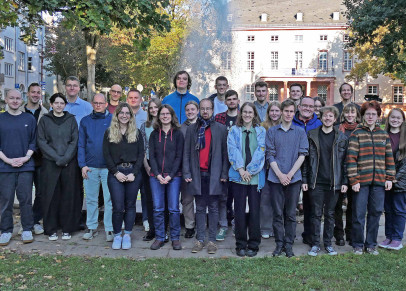 Eine Gruppe Menschen steht in einer Grünanlage. Im Hintergrund sind ein Springbrunnen und ein großes Gebäude zu sehen.