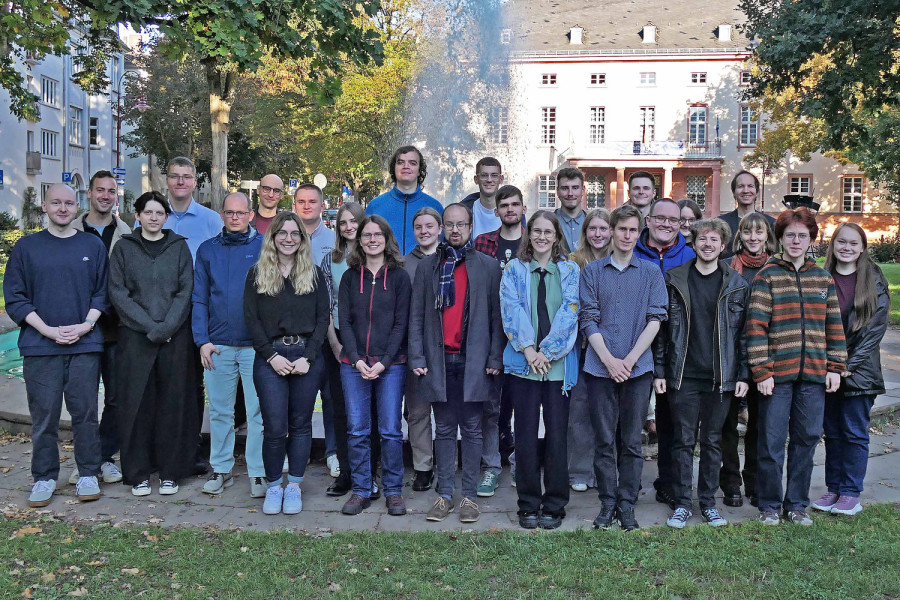 Eine Gruppe Menschen steht in einer Grünanlage. Im Hintergrund sind ein Springbrunnen und ein großes Gebäude zu sehen.