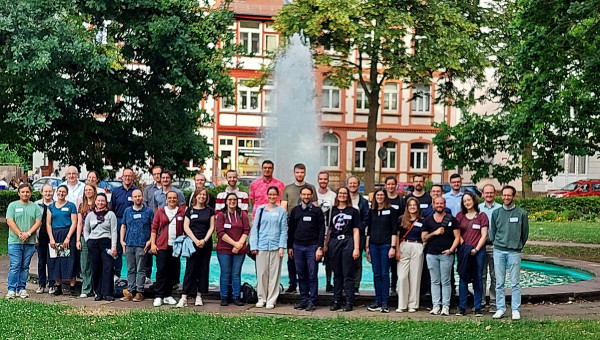 Eine Gruppe Menschen steht vor einem Springbrunnen mit großem Becken und Fontäne auf einem parkartigen Platz.