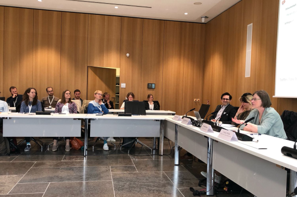 In einem getäfelten Raum sitzen Menschen im Geviert an Tischen. Rechts sieht man teilweise eine Leinwand, auf die ein Beamer eine Präsentation projiziert.